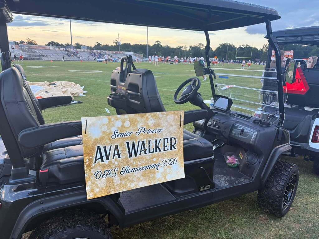 A golf cart with a sign reading "AVA WALKER" parked on a field. Behind it, a stadium is visible.