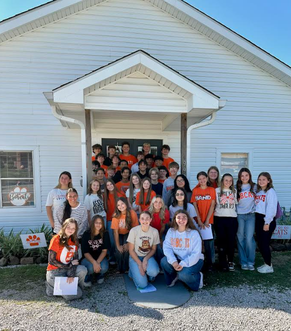 Group of students in orange and white shirts poses outside a white building. Some students are kneeling, others are standing.