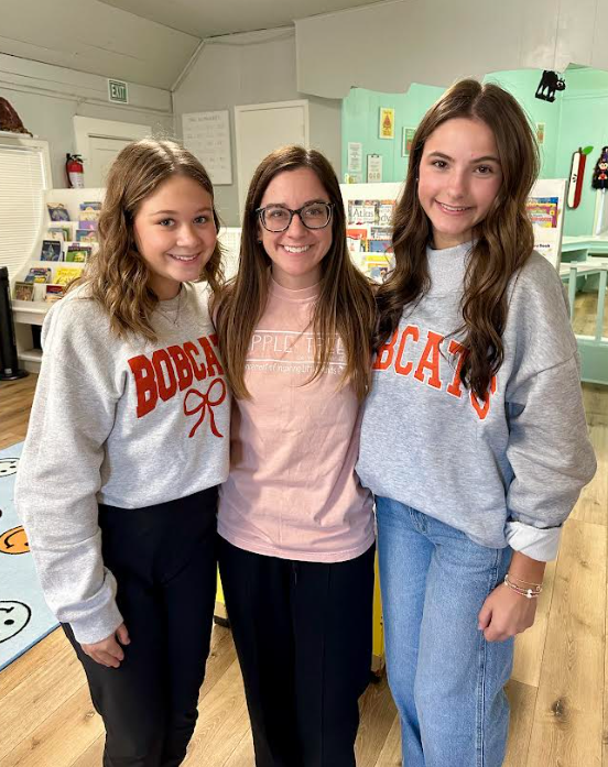 Three young women stand close together in a room, with shelves of books and a rug.
