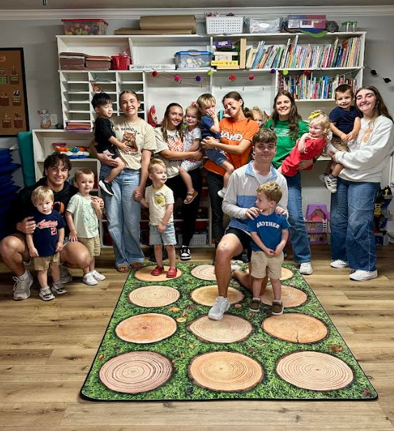 A classroom with wooden floor, bookshelves, toys, and a rug. Group of adults and children gather, some holding kids.