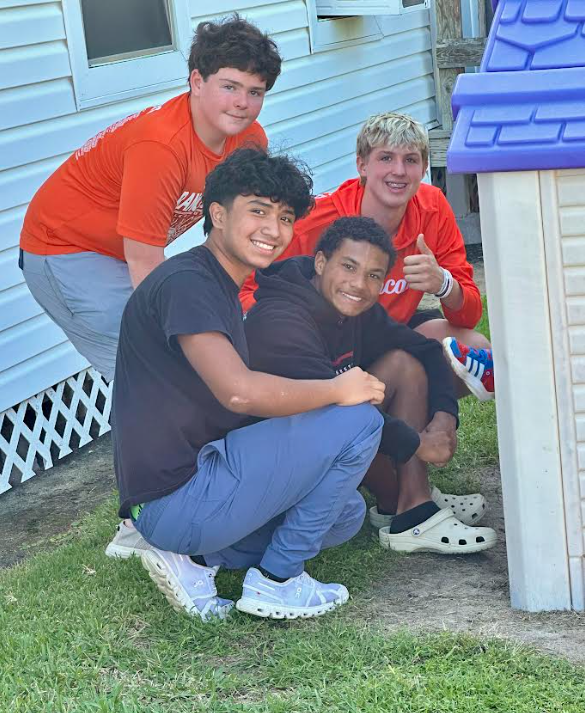 Four boys in orange and black shirts, one kneeling on grass, others posing beside a blue-roofed structure.