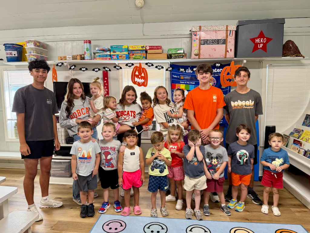 A group of children and adults in a room with a rug, shelves, and a Halloween decoration.