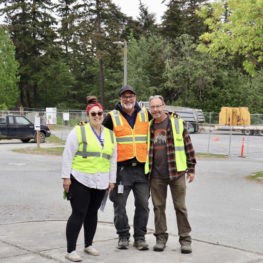 Three people wearing high-visibility safety vests—two men and one woman—pose together and smile in an asphalt parking area. In the background, a chain-link fence surrounds a construction site with various trucks, equipment, and a thick forest of deciduous and evergreen trees.