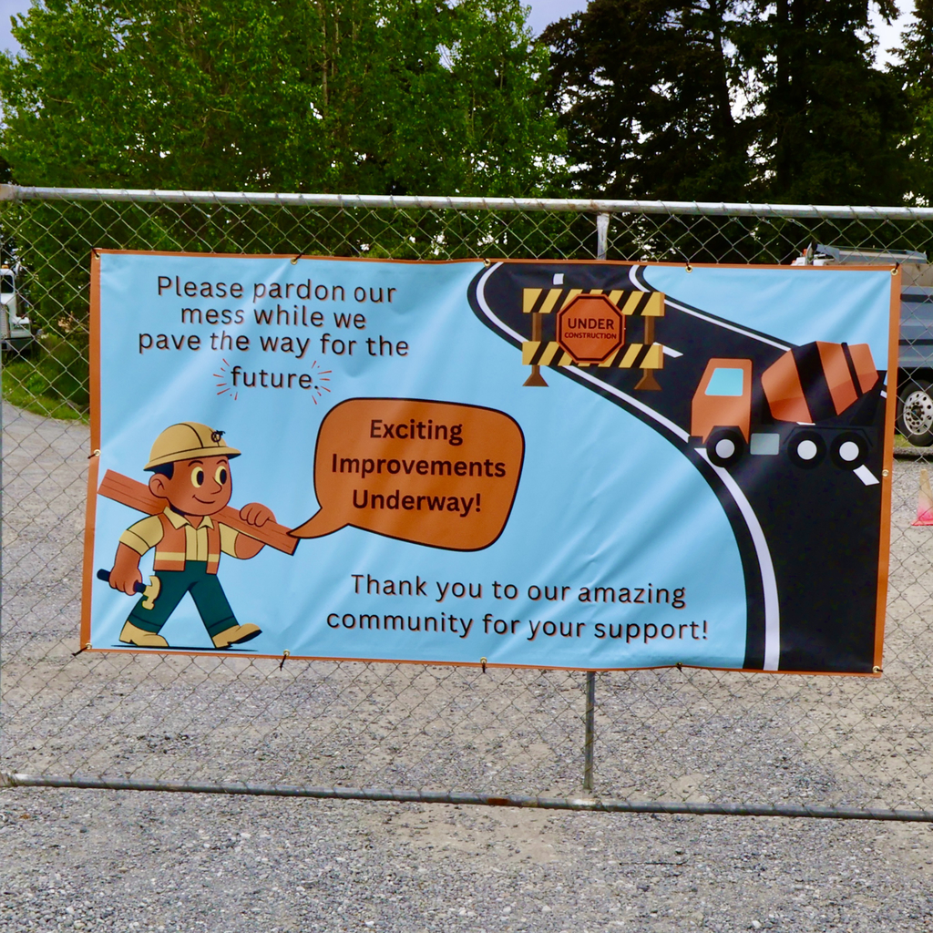 A bright blue construction banner hangs on a chain-link fence, featuring a friendly cartoon construction worker and the text, "Please pardon our mess while we pave the way for the future. Exciting improvements underway."