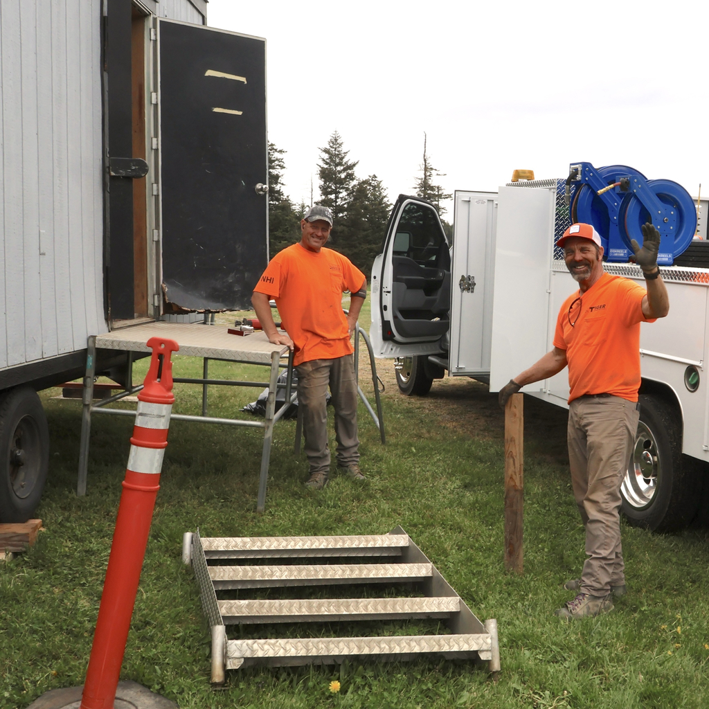 Two construction workers in bright orange t-shirts smile near a white service truck and a grey mobile office trailer. One man stands on the trailer's metal steps while the other stands on the grass, waving to the camera next to a set of detached metal stairs and an orange safety pylon
