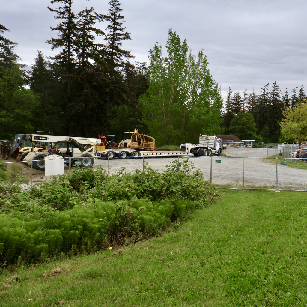 A wide shot of a construction staging area enclosed by a chain-link fence, featuring heavy equipment including a yellow bulldozer on a flatbed trailer and a telehandler. A paved parking lot with several cars is visible to the right, all set against a dense backdrop of tall evergreen trees under an overcast sky