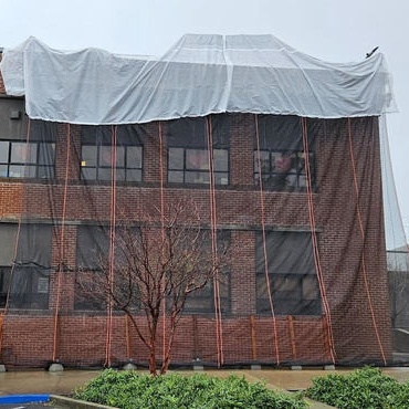 The brick elementary school Milton building with tarps stretched across the top, and debri catching mesh curtains covering the side from top to bottom.