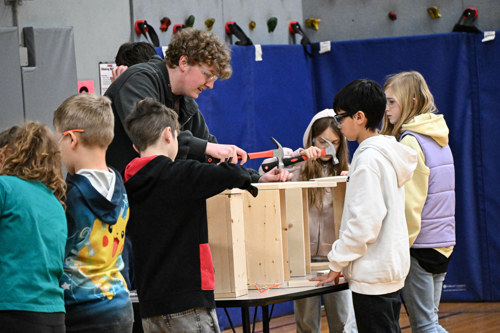 An older student mentor guides a group of elementary-aged students in a hands-on woodworking activity in a school gymnasium. Several children gather around a table, using hammers to assemble a wooden structure that resembles a birdhouse or small shelf. Some students wear safety glasses. A climbing wall is visible in the background.