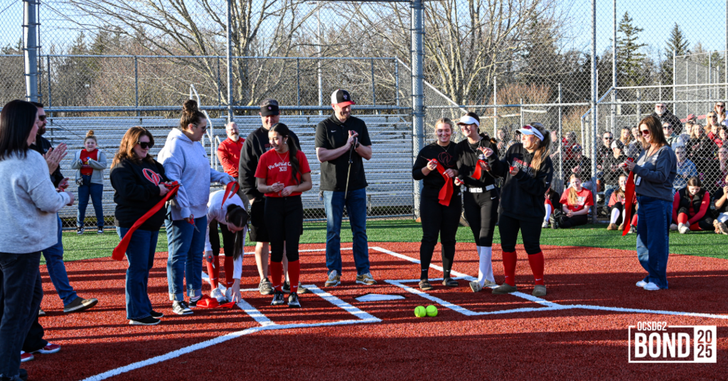 Softball team members, district staff members, and school board members smiling holding cut ribbon.