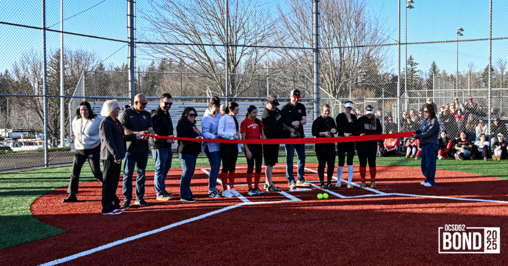 Softball team members, district staff members, and school board members smiling poised to cut ribbon. 