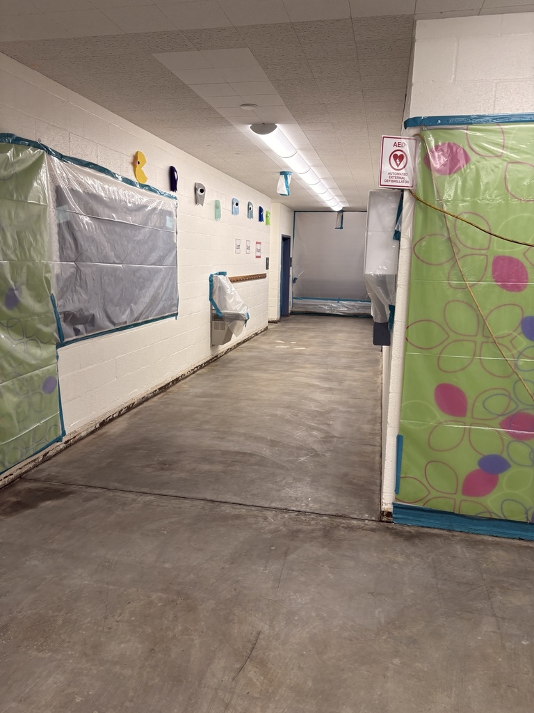 Second view of Holcomb hallway showing classroom doorways protected with plastic sheeting and blue tape. Colorful bulletin board decorations are visible beneath the protective covering.
