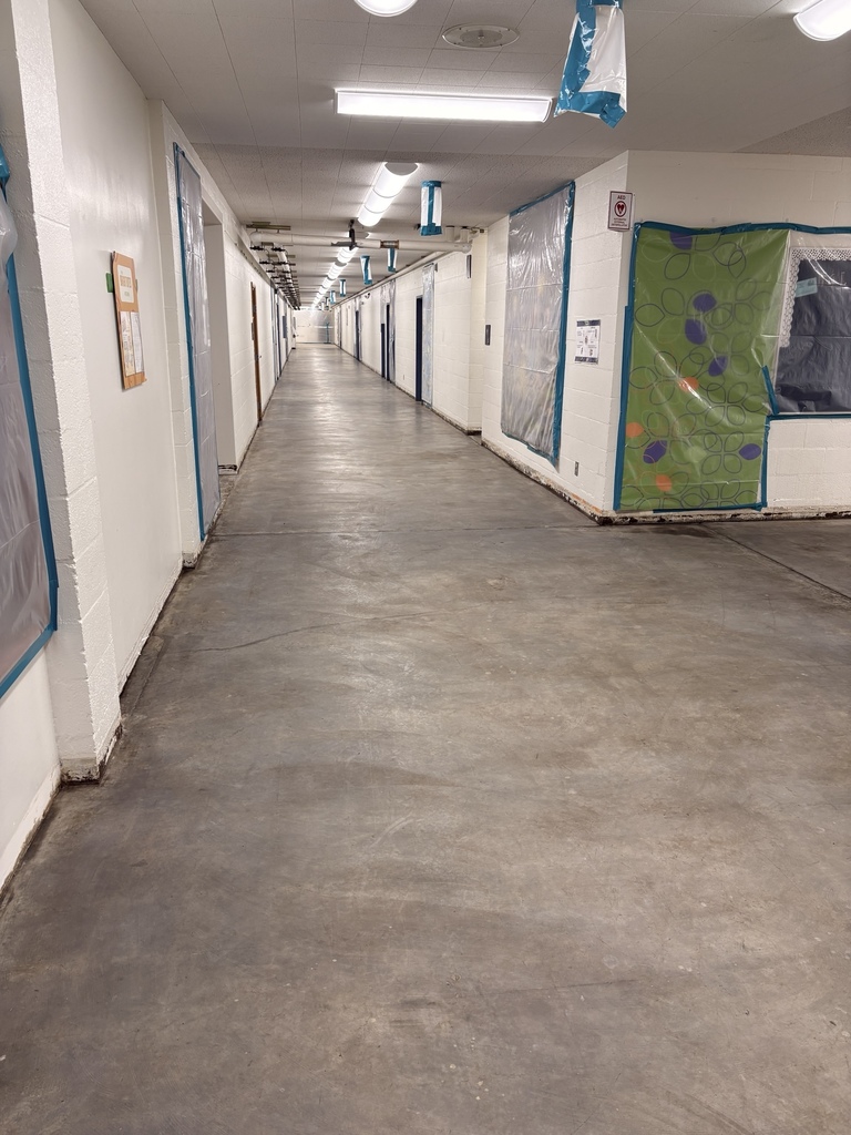 Holcomb Elementary hallway with floor tiles removed during abatement work. White walls line the corridor, classroom doors and bulletin boards are covered with protective plastic sheeting sealed with blue tape, and air filtration equipment hangs from the ceiling.