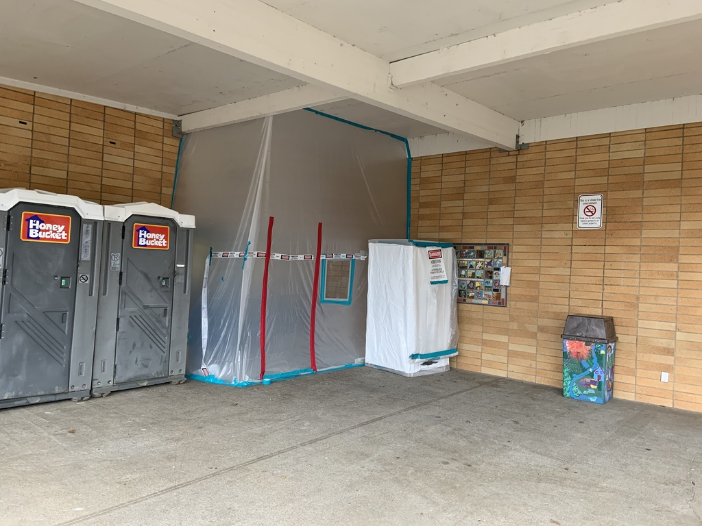 Holcomb gymnasium or multipurpose room showing protective barriers sealed with blue and red tape. Two portable restrooms are positioned in the space, with wood-paneled walls visible in the background.