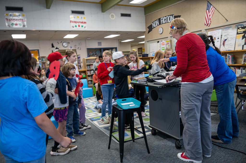 Students gathered in a library listening to a teacher who is standing at a podium up front