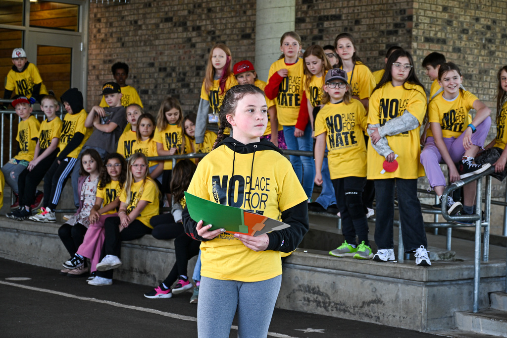 A girl holding folders standing in front of a group of students wearing yellow shirts that say "No Place for Hate"