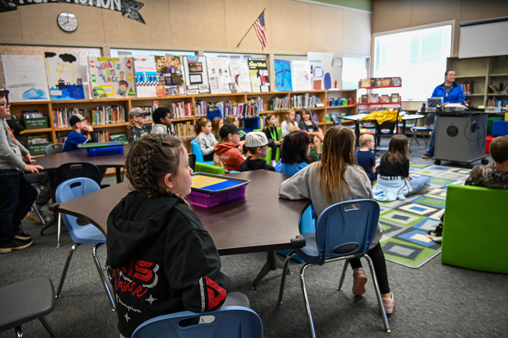 Students gathered in a library listening to a teacher who is standing at a podium up front