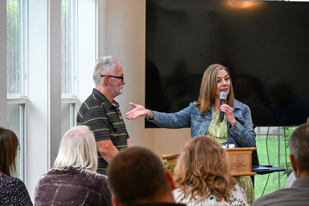 Woman, smiling, holding a microphone and gesturing with an outstretched arm to a man standing nearby