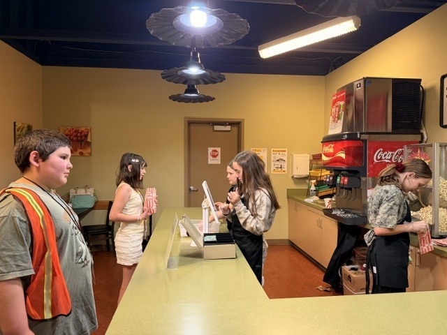 Students work behind and in front of a snack bar counter with a Coca-Cola fountain machine and popcorn maker, with one student appearing to process a transaction on a point-of-sale tablet.