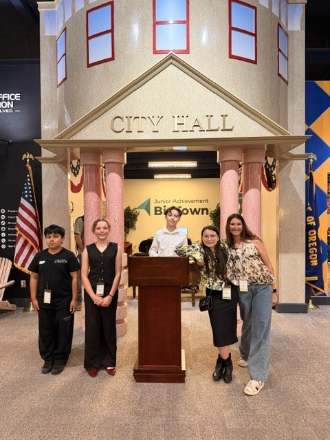 Four students stand together in front of the Junior Achievement BizTown City Hall façade, which features columns, an American flag, and the Oregon state flag.