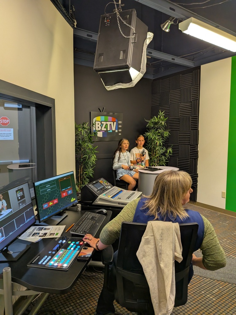 Two students sit at an anchor desk inside a simulated BZTV television studio while an adult volunteer operates a production control board in the foreground, with studio lights and a green screen visible.