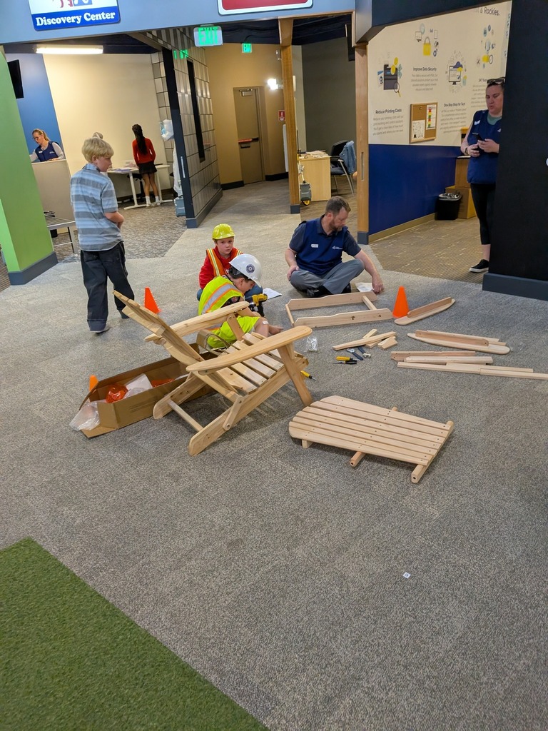 Two students wearing hard hats and safety vests assemble a wooden Adirondack chair on the floor of a career exploration center, assisted by an adult volunteer, with orange safety cones marking the work zone.