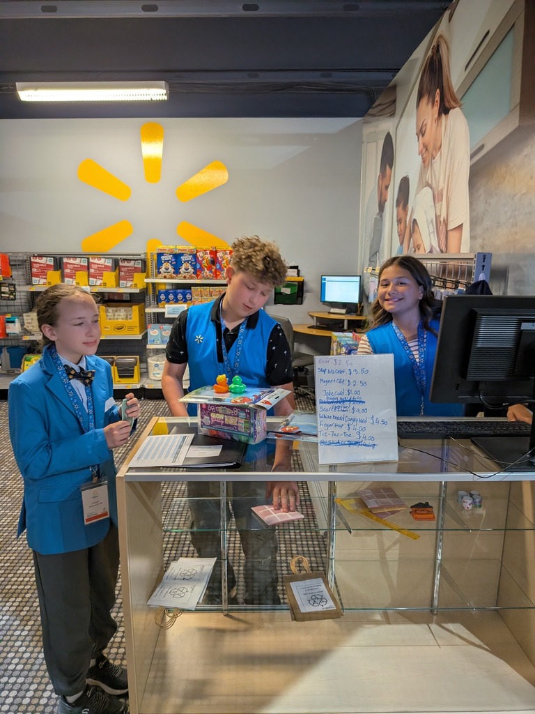 Three elementary school students in blue Walmart vests stand behind a checkout counter at a simulated Walmart store, with product shelves and a handwritten price list visible in the background.