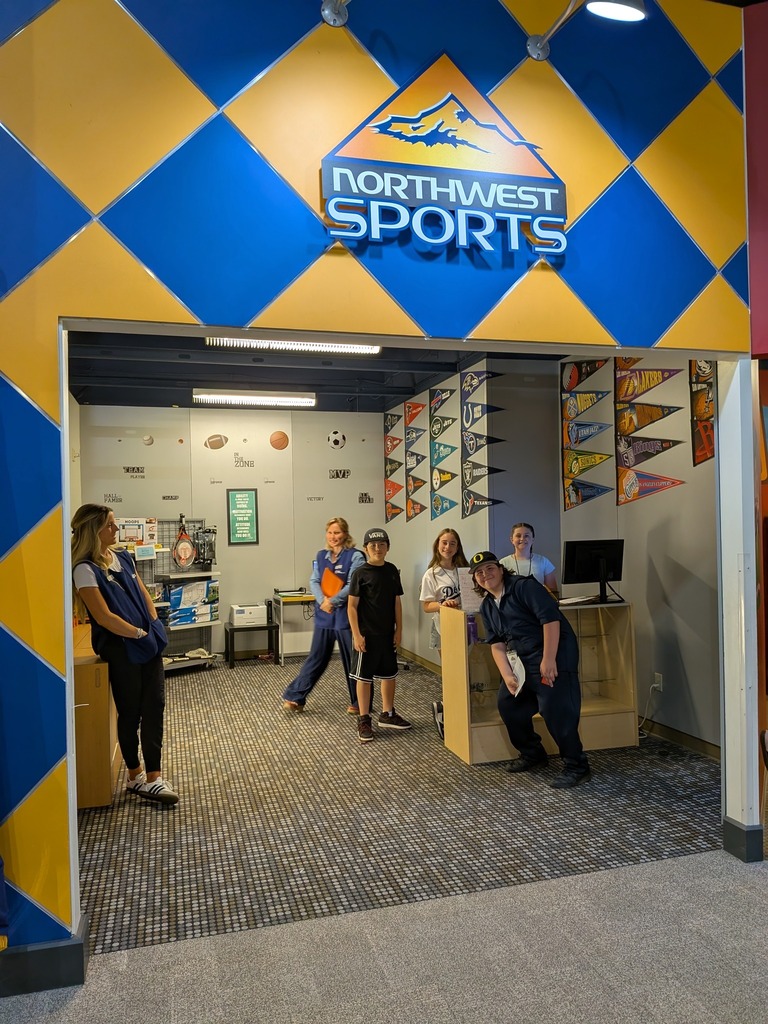 A group of students and adult volunteers pose inside a simulated Northwest Sports store, surrounded by team pennants and sports-themed décor on a blue and gold diamond-patterned wall.