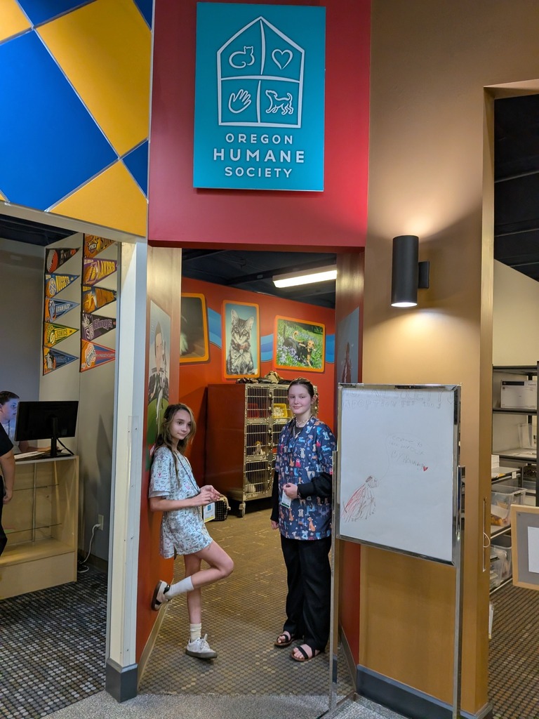 Two students stand in the entrance of a simulated Oregon Humane Society exhibit, featuring orange walls with animal photography murals and a wire kennel holding a stuffed animal inside.