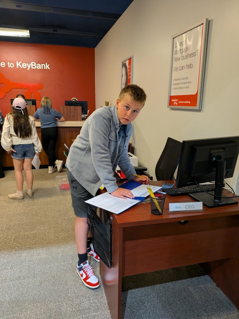 A student leans over the CEO desk at a simulated KeyBank branch, reviewing paperwork, with other students visible at teller windows in the background.
