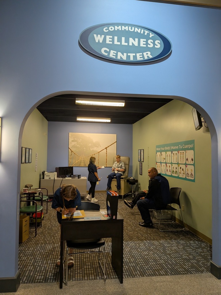 Students and adult volunteers work inside a simulated Community Wellness Center, with one student writing at a front desk, another seated in an exam chair, and a man waiting in a chair nearby.