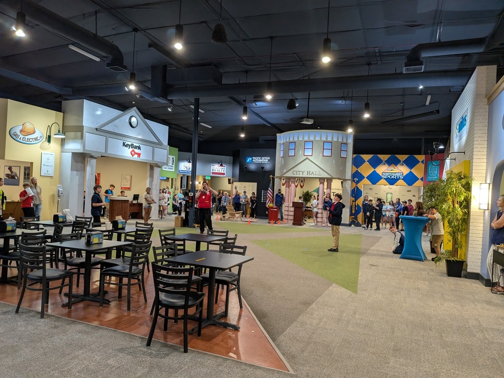 Wide-angle view of the full Junior Achievement BizTown facility, showing the miniature city layout with storefronts including KeyBank, City Hall, Northwest Sports, and Gas & Electric, with students and chaperones filling the space.