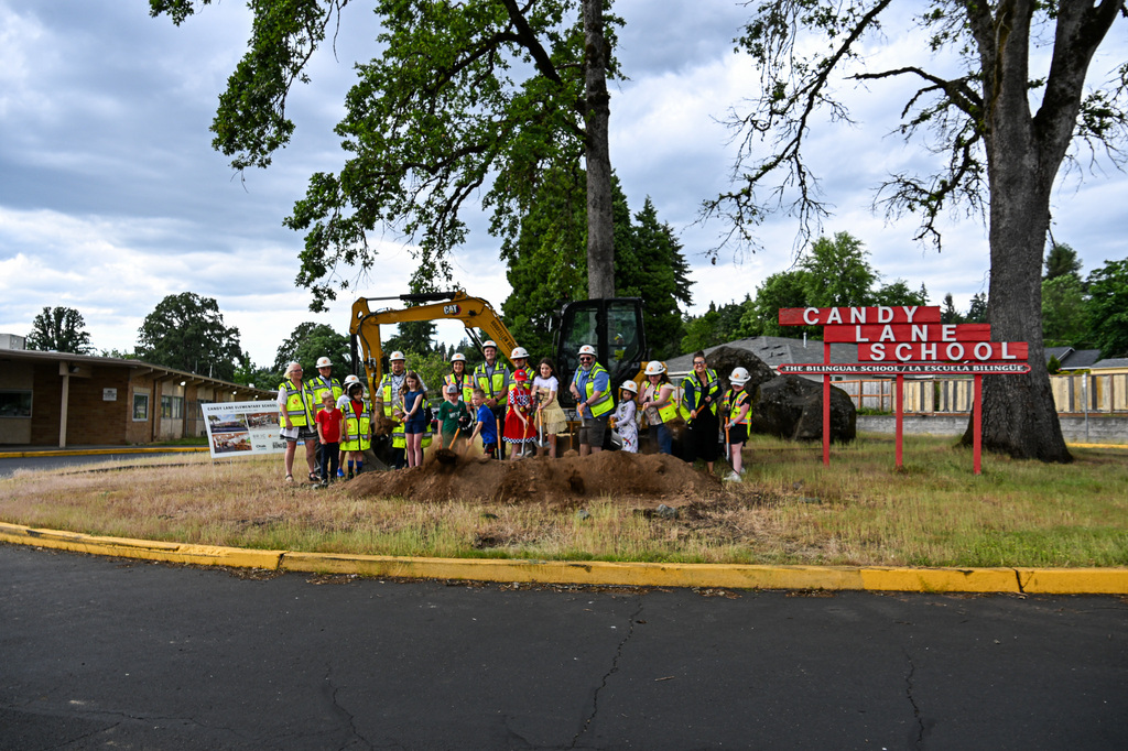A group of Candy Lane students, families, and bond team members stand with ceremonial shovels at the groundbreaking site beside the school's red bilingual sign.