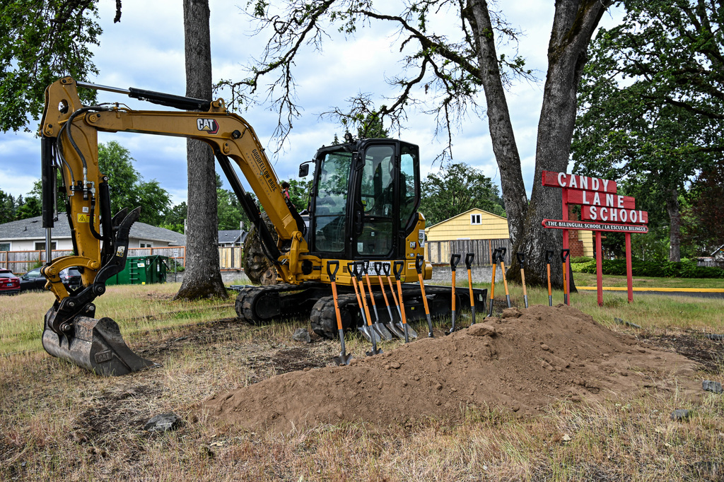 A yellow CAT excavator and a row of ceremonial shovels rest in a fresh mound of dirt at the Candy Lane construction site, next to the school's red bilingual sign and a large oak tree.