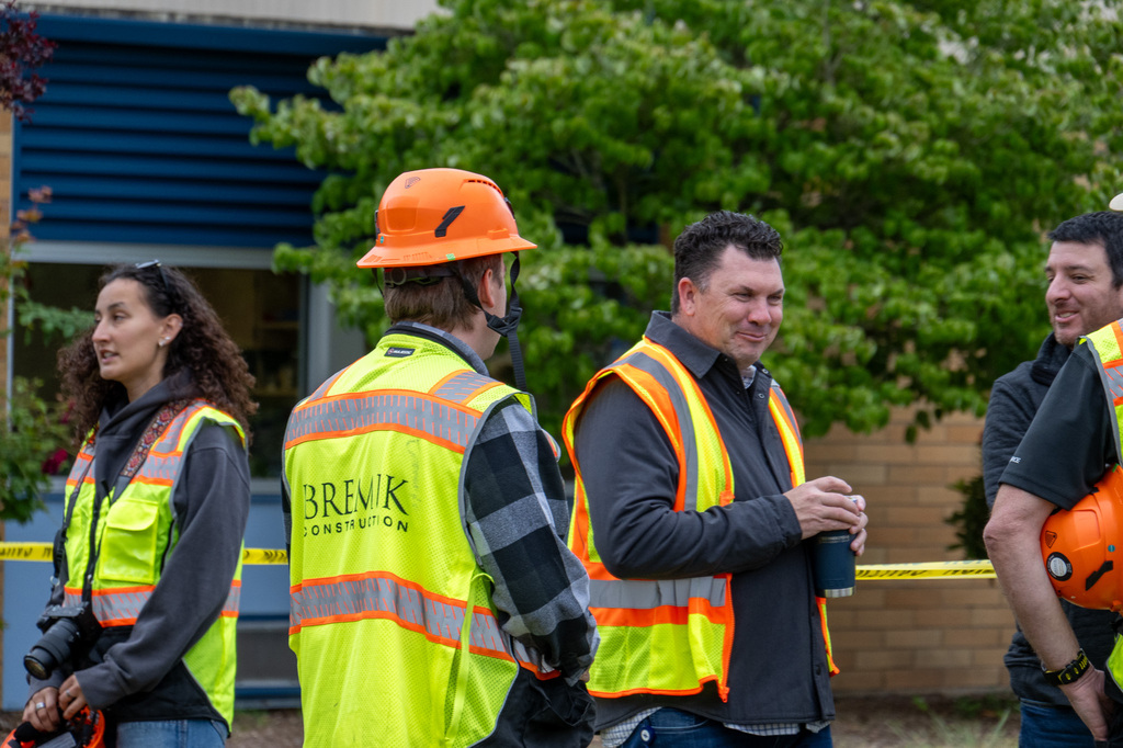 Individuals in yellow safety vests and orange hard hats stand in a group smiling and talking