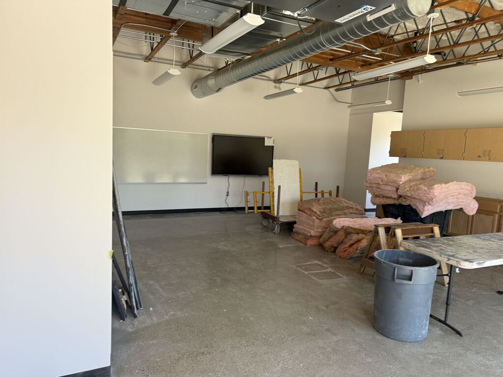 A room under construction with insulation laying on the ground. A white dry erase board and monitor have been installed on the wall.