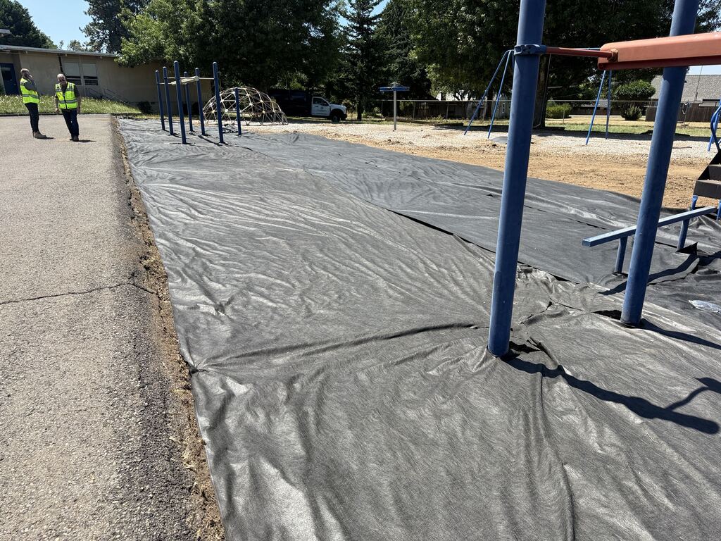 A playground with black plastic laid down getting ready for bark chips to be delivered. Two people standing in the background wearing safety vests and hard hats.