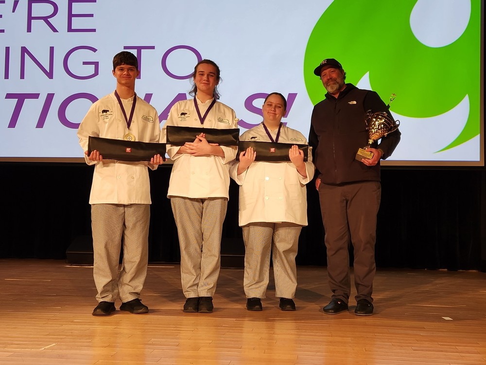 Three students dressed as chefs holding knife sets and teacher with trophy