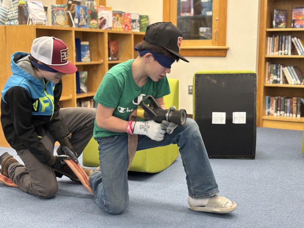 Two male students hold a fire house and have their blindfolds one while moving on one knee while they go to rescue a person in a "smoke-filled building"