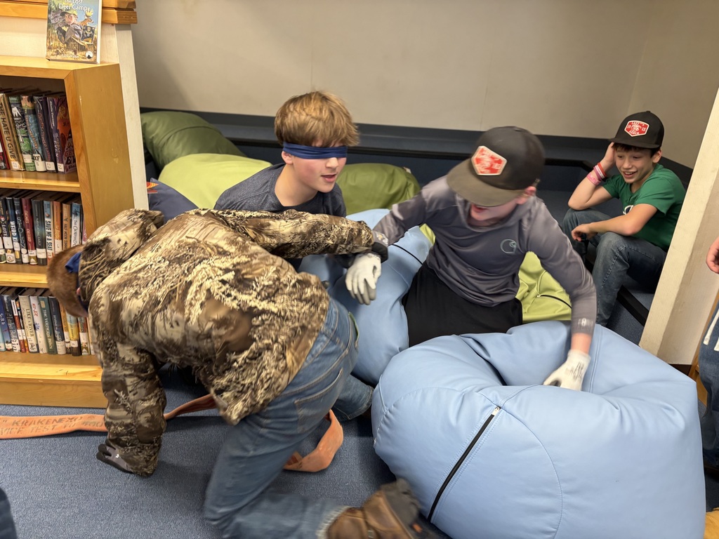 Two male students hold a fire house and have their blindfolds one while moving on one knee while they go to rescue a person in a "smoke-filled building"