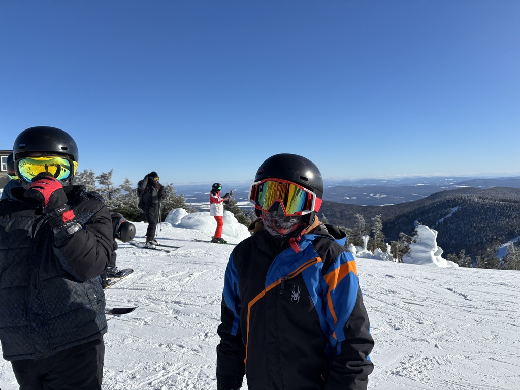 Two snowboarders decked in their gear pose at the top of the mountain at Jay Peak