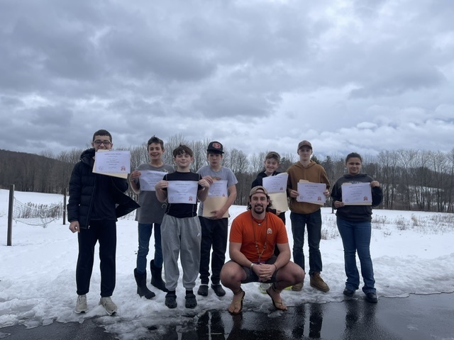 Devin squatting on the ice while the outdoor wilderness group stands in the snow holding their certificates 