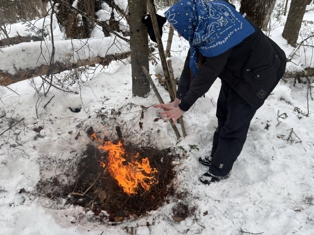 A student wearing a giant bandana and warming her hands around a fire built outside 