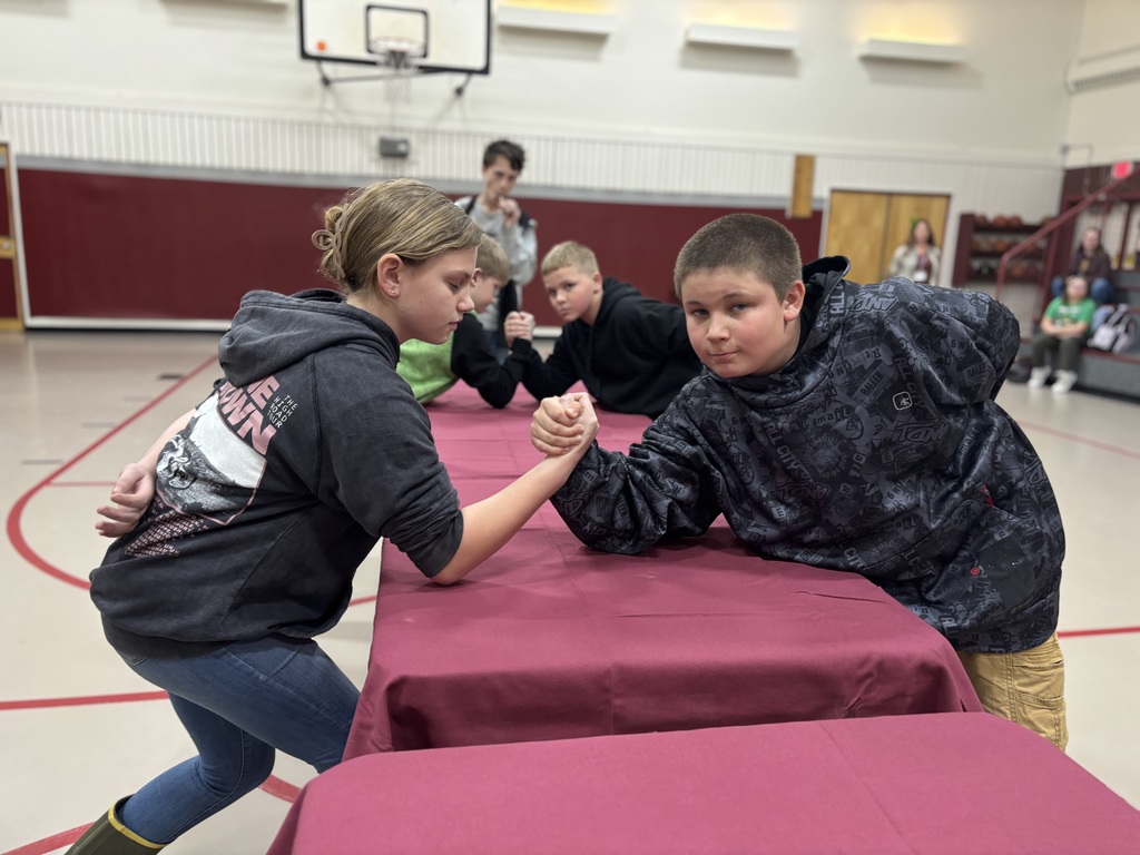 A 5th grade and 6th grade student are arm wrestling on a table in the gym. 
