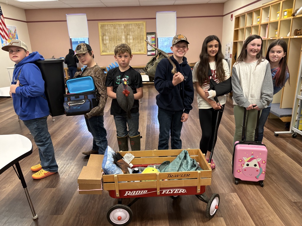 a group of 5th graders hold various items (wagon, suitcase, trashcan, lunch box, bag on a stick, and turkey)