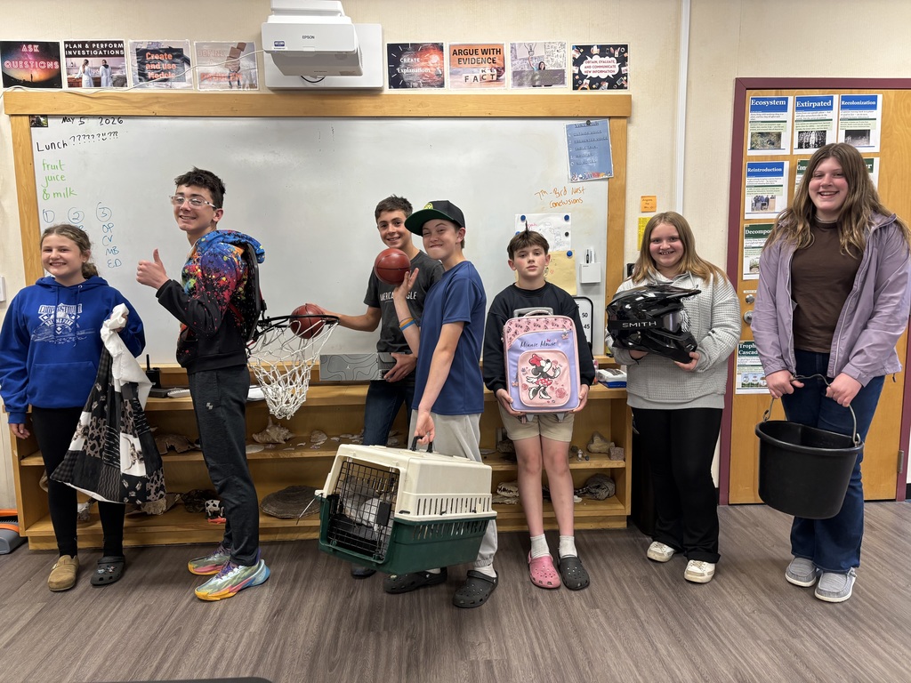 7th grades stand in a line holding various objects (a quilt bag, a basketbal hoop, a shoebox, cat carrier, suitcase, dirtback helmet, and a horse bucket)