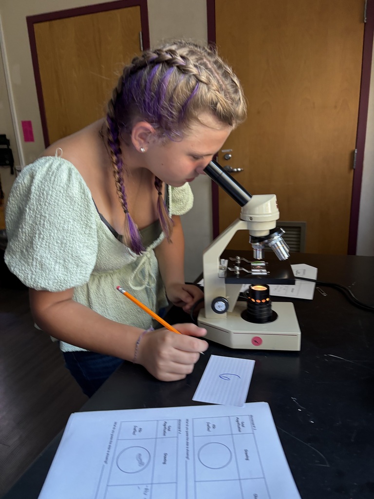 a 6th grade girl peers through a microscope and holds a pencil 