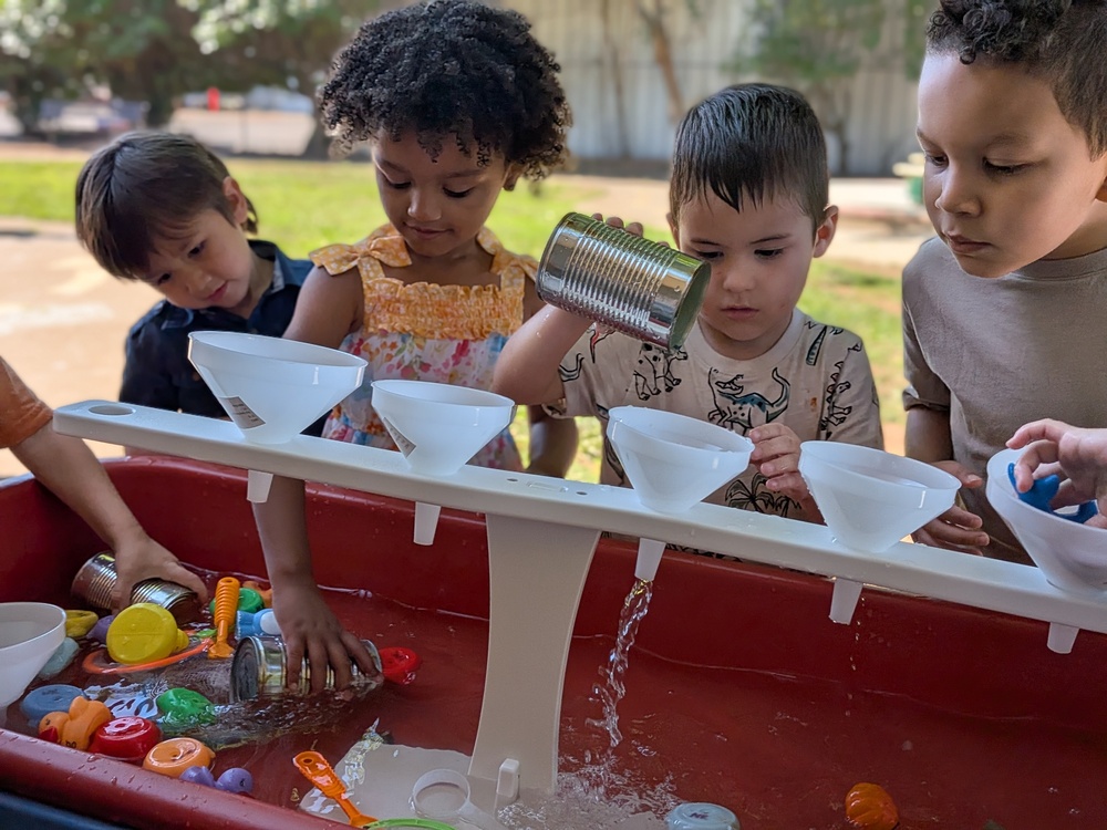 Four Preschool Children standing at a water table looking, pouring and scooping water through 5 funnels on a stand.