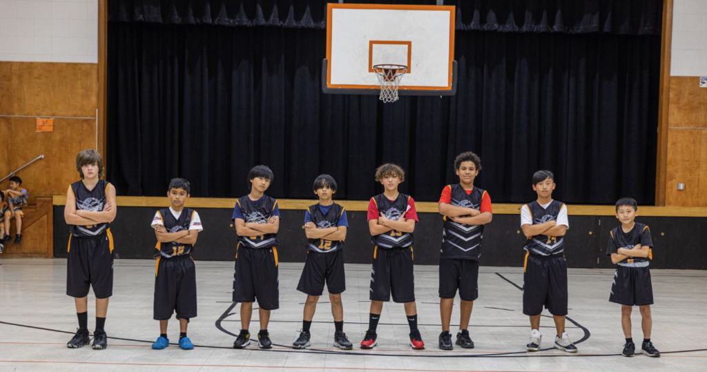 "The Studios boys basketball team posing together on a court. Eight players stand in a line with their arms crossed, wearing black, grey, and orange 'The Studios' jerseys. They are positioned in front of a black-curtained stage under a basketball hoop, looking focused and determined."