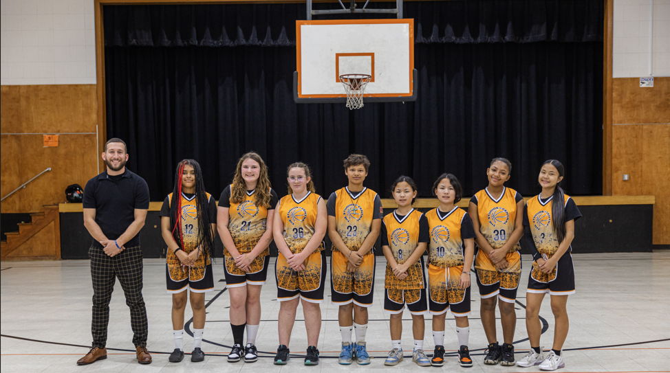 "A professional team photo of the Studios girls basketball team standing on a gym court with their coach. The eight players are wearing matching orange, black, and white jerseys with 'The Studios Central' logo. They are lined up in front of a stage with black curtains, positioned directly under a basketball hoop."
