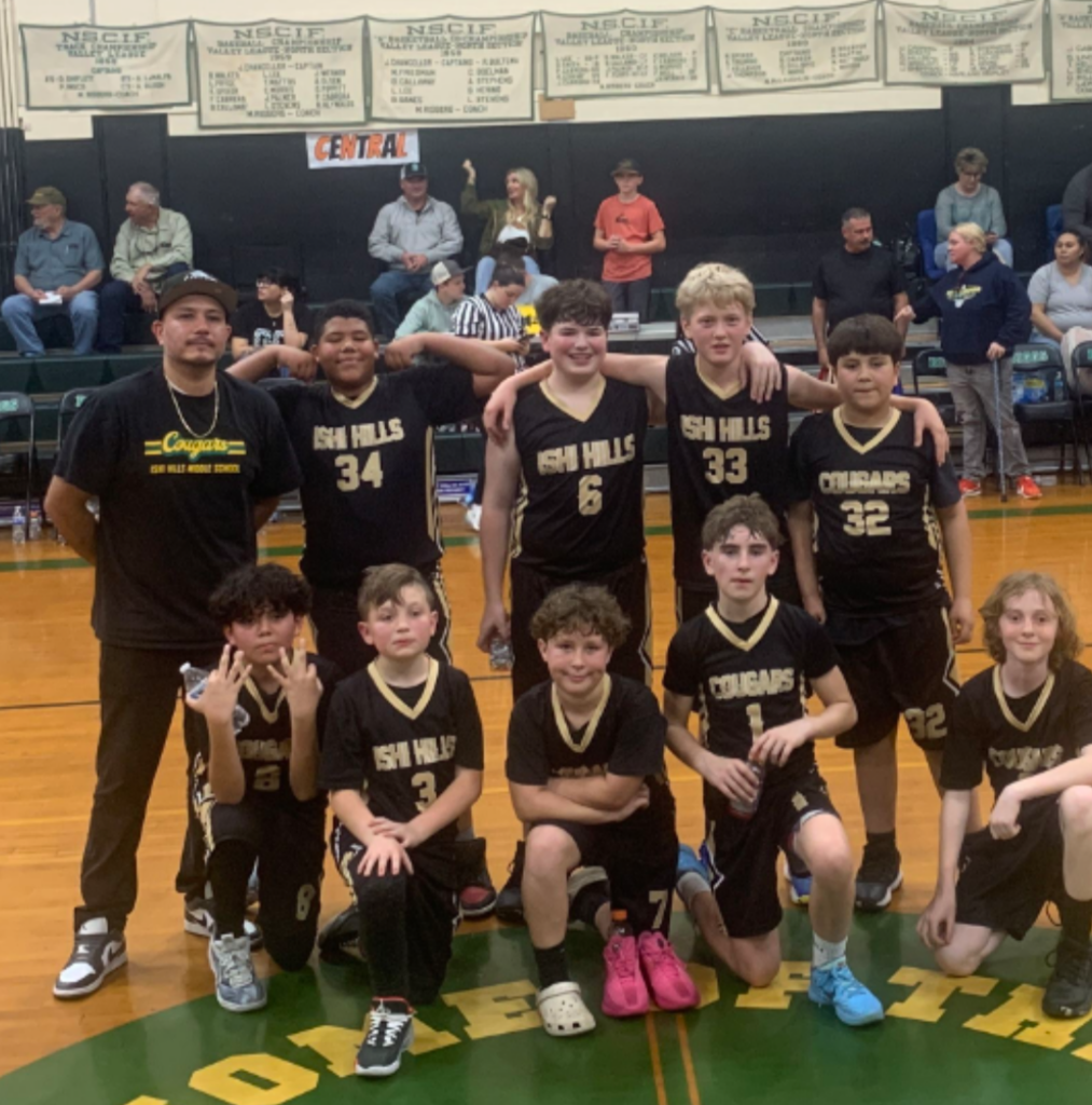 "The Ishi Hills 6th grade boys basketball team posing with their coach on a gymnasium court. The team is dressed in black and gold 'Ishi Hills' and 'Cougars' jerseys. Some players are kneeling in the front row while others stand behind them with their arms around each other, celebrating their second-place finish."